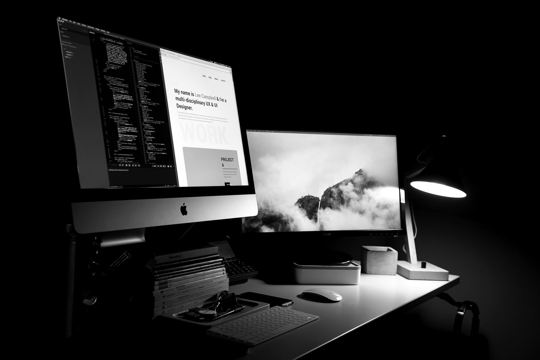 A designer's desk in a dark room: one screen with a website's design and code, another beside it under a lamp