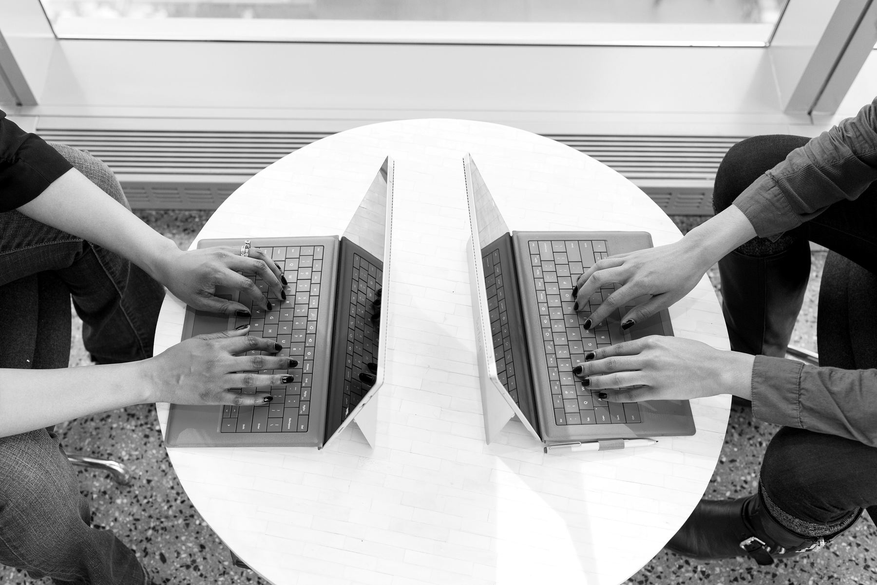 Two people working opposite each other at a round table, each typing on a laptop
