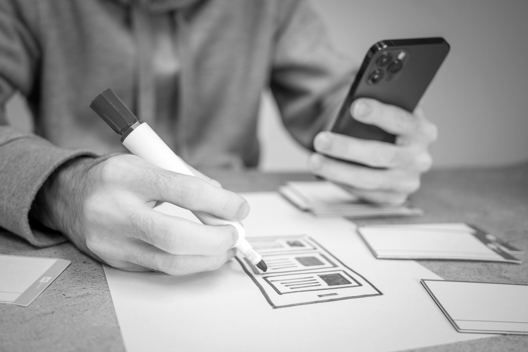 A hand drawing a website's page layouts on paper, the other holding a phone to try the design on